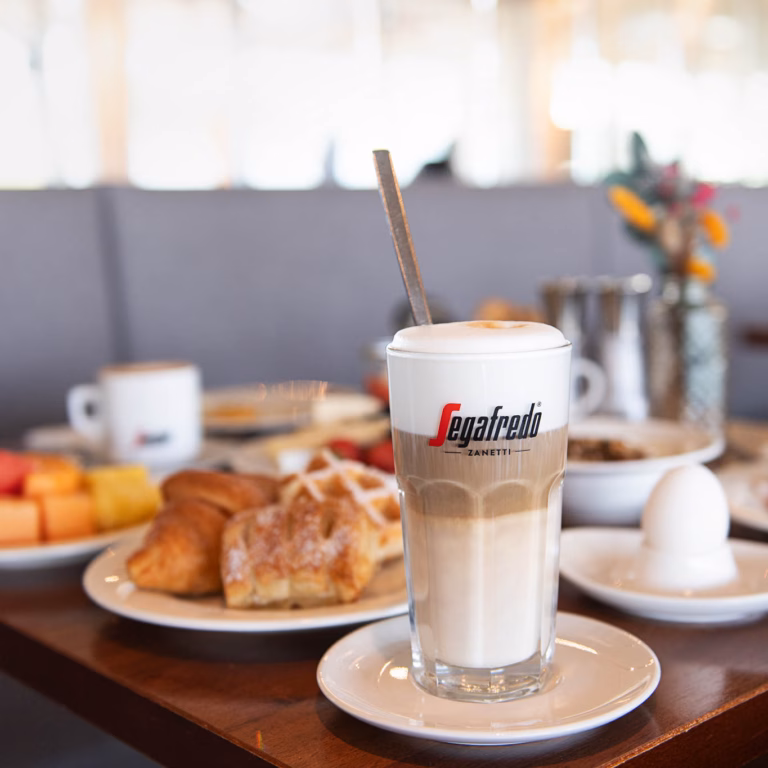Imagebild Frühstückstisch mit einem Glas Milchkaffee und Gebäck im Hintergrund