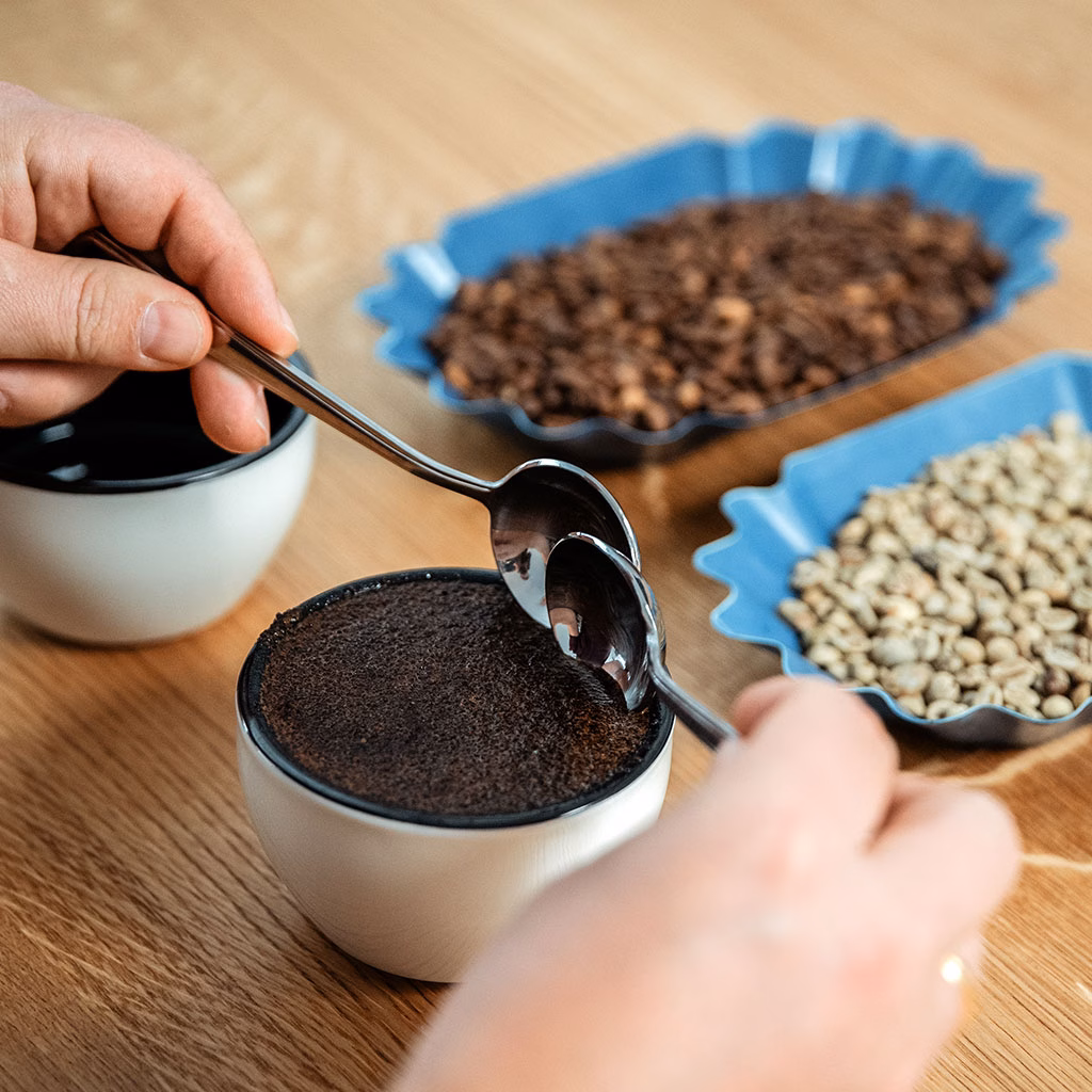 Zwei Hände fahren mit Löffeln in eine Schüssel, im Hintergrund einige Kaffeebohnen