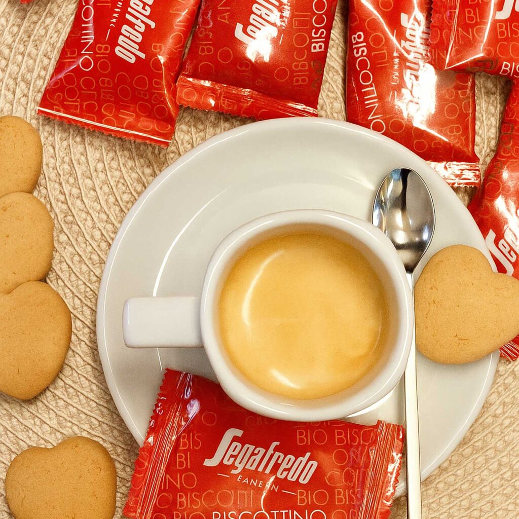 Moodbild von oben mit Kaffeetasse auf einer Untertasse mit Löffel und Kaffee darin, Bio-Kekse in Herzform ausgepackt und in der Verpackung